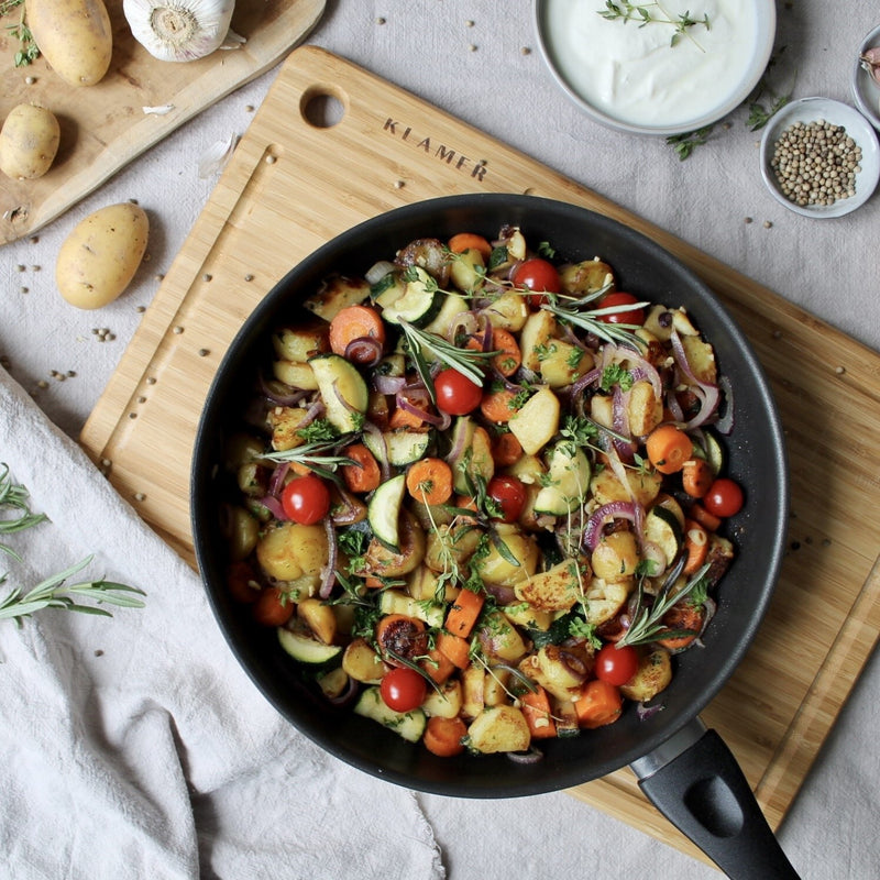 Gemüsepfanne in Profi Edelstahl-Bratpfanne 28cm mit Kartoffeln, Zucchini, Tomaten und Kräutern auf Holzunterlage.