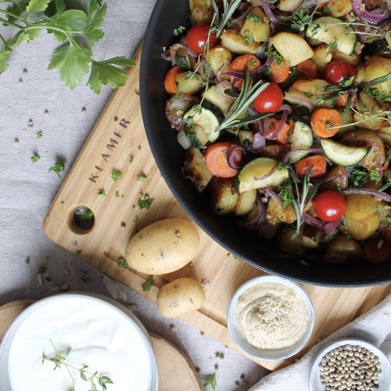 Profi Edelstahl-Bratpfanne 28cm mit gebratenem Gemüse und Kräutern auf einem Holzbrett.