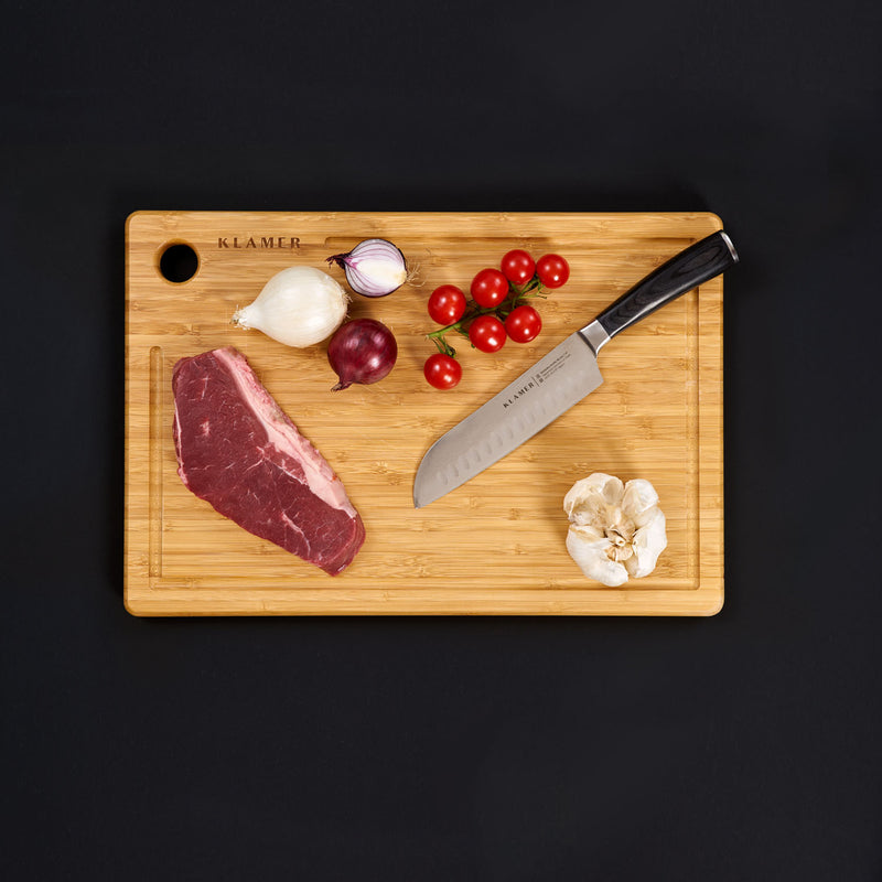 Ein Santoku Damastmesser auf einem Holzschneidebrett mit Fleisch, Zwiebeln, Tomaten und Knoblauch.